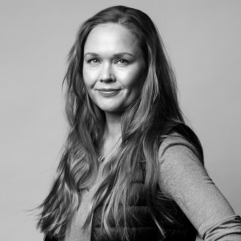 Pitkähiuksinen nainen, jolla on pitkähihainen paita ja liivi, seisoo kameraan päin tavallista taustaa vasten.