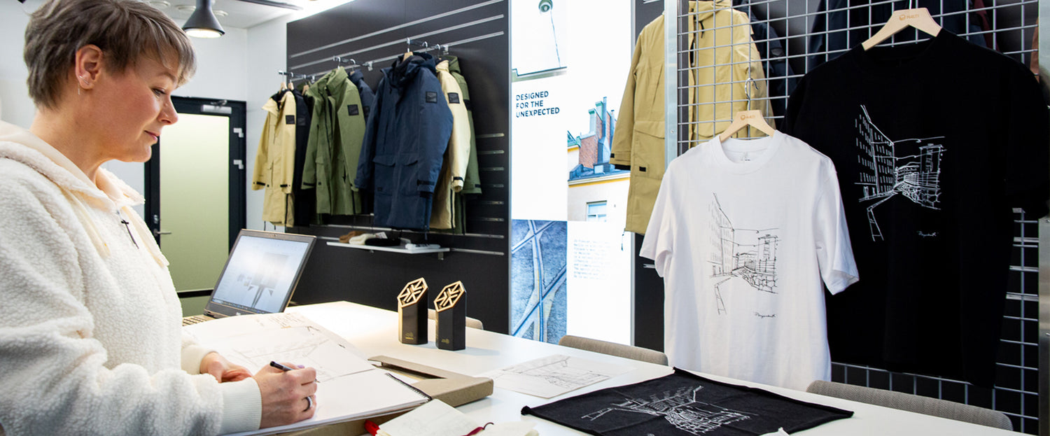 Henkilö piirtää pöydän ääressä, jossa on takkeja ja graafisia t-paitoja ripustimissa ja pöytä lähellä.