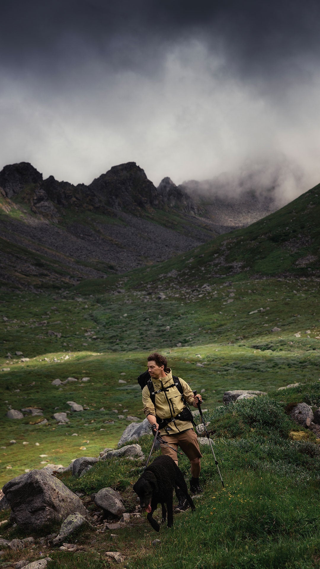 Henkilö, jolla on vaellussauvat ja reppu, kävelee mustan koiran kanssa kallioisessa vuoristomaisemassa pilvisen taivaan alla.