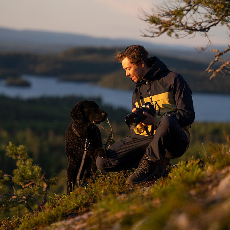 Ulkovaatteisiin pukeutunut mies polvistuu rinteessä kameran kanssa, vastassa musta koira hihnassa, taustalla järvi.