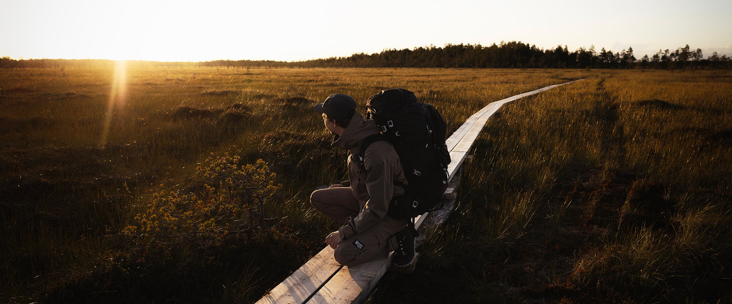 Henkilö, jolla on iso reppu mukanaan, kyyristelee puupolulla ruohokentän halki auringonlaskun aikaan.