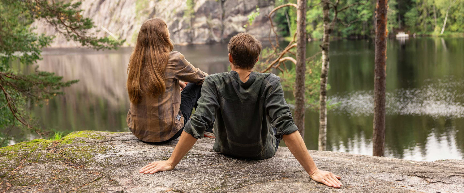Kaksi ihmistä istuu kalliolla rauhallisen järven rannalla, kasvot poispäin, puiden ja metsäisten kallioiden ympäröimänä.