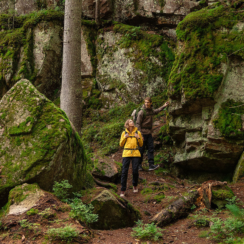Kaksi ihmistä seisoo metsänpohjalla sammaloituneiden kivien ja puiden vieressä, katselee ylöspäin ja tarkkailee ympäristöään.
