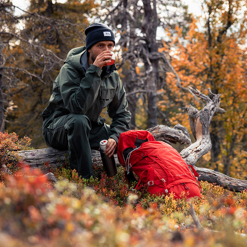Ulkoiluvarusteisiin pukeutunut henkilö istuu tukin päällä, juo kupista, punainen reppu ja termoskannu lähellä metsässä.