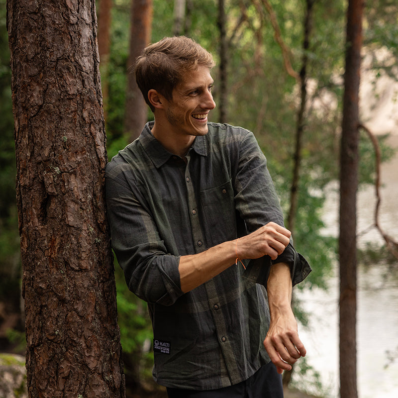 Mies seisoo ulkona puun vieressä ja käärii hihansa ylös, taustalla metsä ja vesistö.