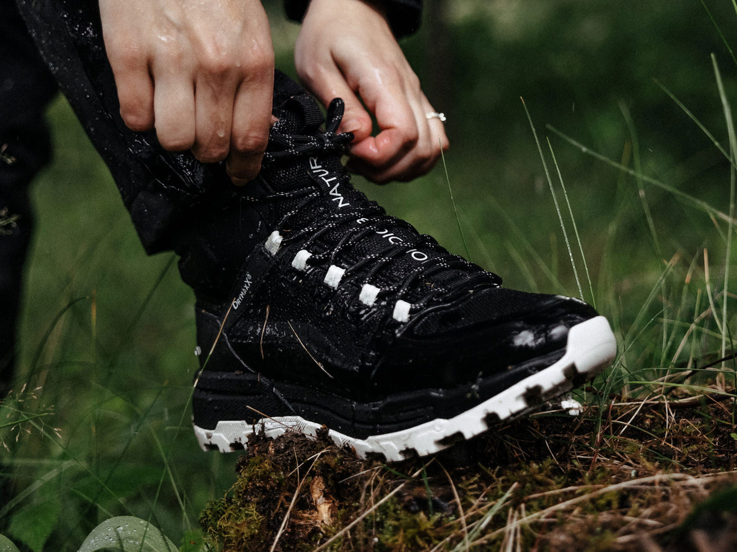Henkilö sitoo mustavalkoisen vaelluskengän mustia nauhoja ulkona märällä ruoholla ja maaperällä.