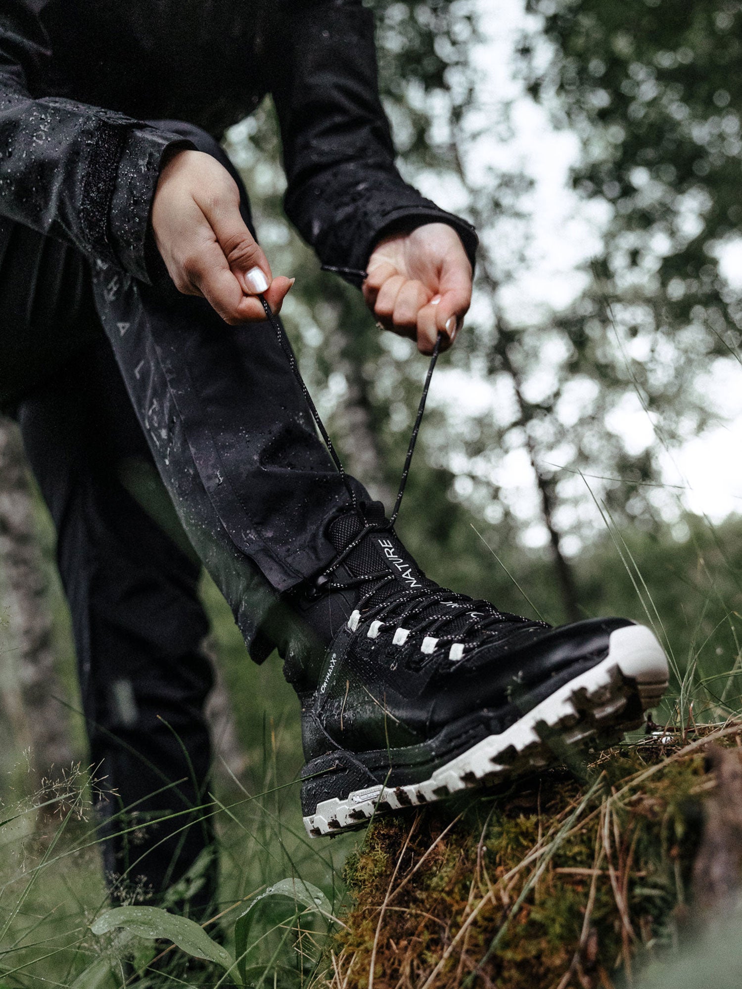 Tummiin vaatteisiin pukeutunut henkilö sitoo mustan vaelluskengän nauhat seisoessaan sammaloituneella metsätukilla.