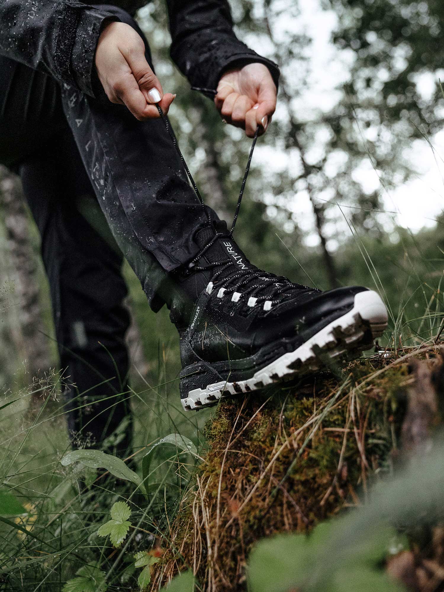 Mustiin ulkoiluvaatteisiin pukeutunut henkilö sitoo mustan vaelluskengän nauhat seisoessaan sammaloituneella tukilla metsässä.