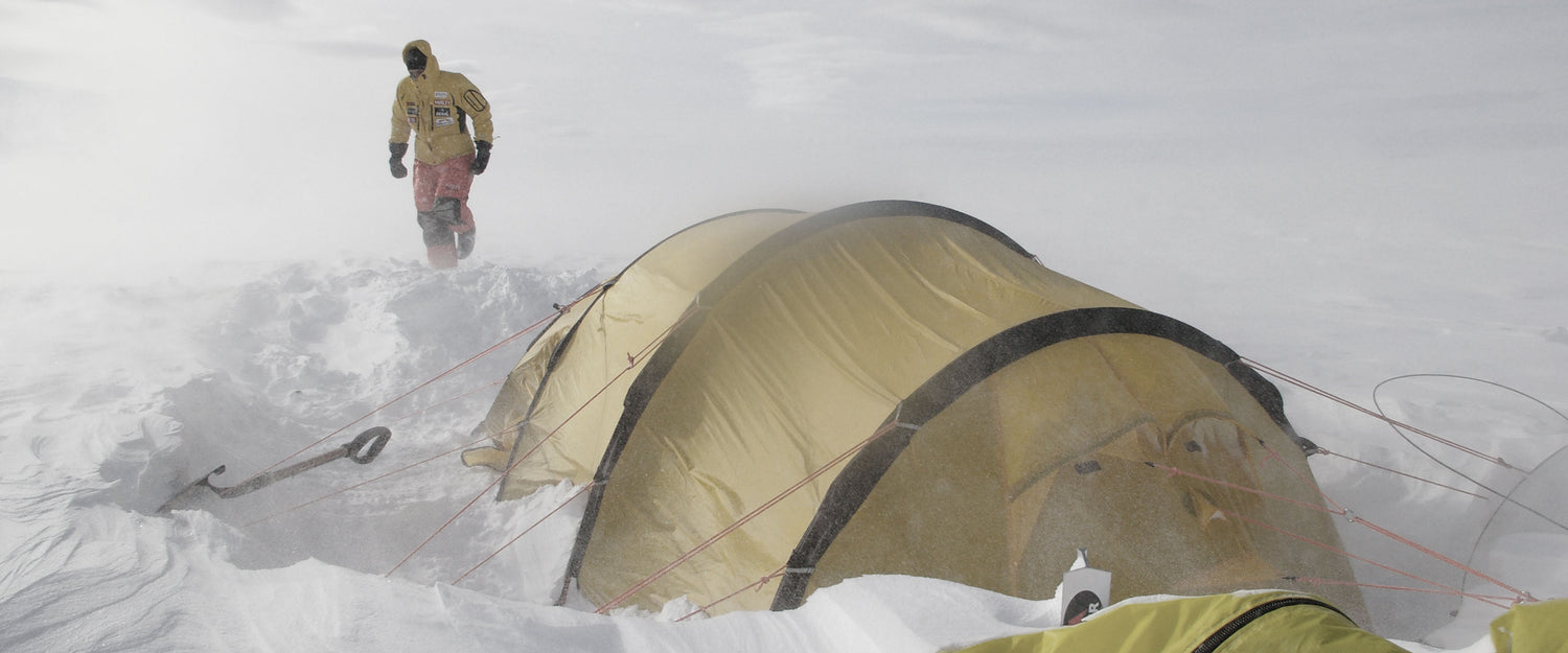 Kylmävarusteisiin pukeutunut henkilö seisoo keltaisen teltan vieressä lumisessa, tuulisessa ympäristössä.