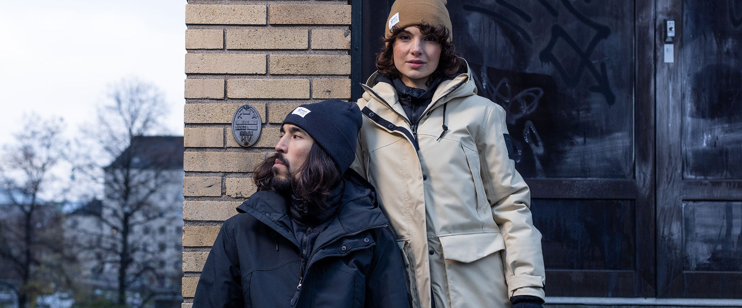 Kaksi talvitakkiin ja pipoon pukeutunutta ihmistä seisoo ulkona tiiliseinän ja graffitilla koristellun oven vieressä.