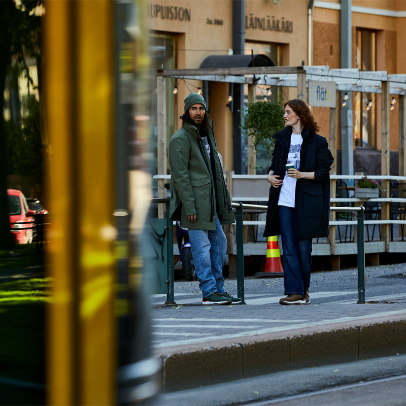 Kaksi ihmistä seisoo kaupungin jalkakäytävällä kaiteen vieressä, ja etualalla kulkee sumea keltainen ajoneuvo.