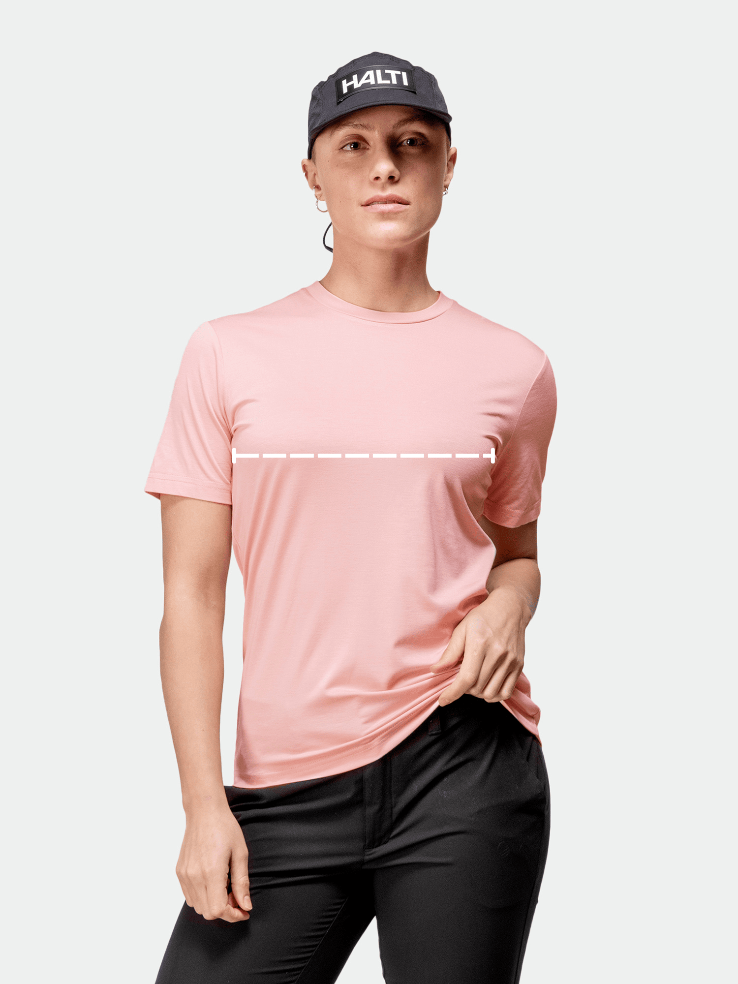Henkilö, jolla on vaaleanpunainen t-paita, mustat housut ja musta HALTI-lippis, pitää toisella kädellä kiinni t-paidan helmasta.