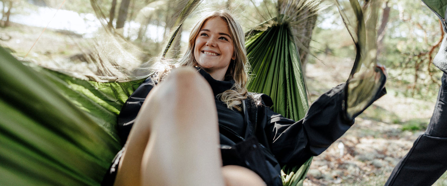 Nainen istuu vihreässä riippumatossa ulkona hymyillen, taustalla näkyy puita ja auringonvaloa.