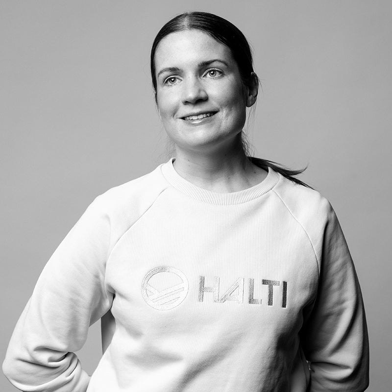 Nainen, jolla on tummat hiukset poninhännässä, yllään vaalea HALTI-verryttelypaita, seisoo tavallista taustaa vasten.