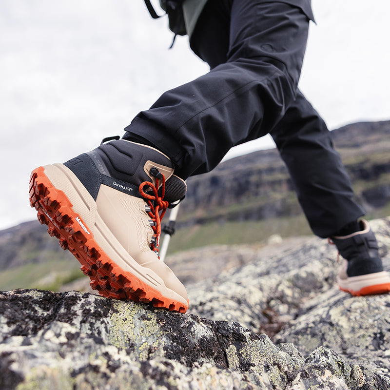 Lähikuva henkilöstä, jolla on vaelluskengät ja mustat housut, kävelee kivikkoisessa maastossa ulkona.