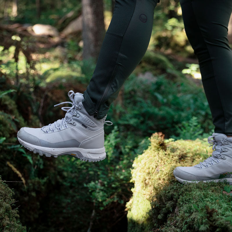 Henkilö, jolla on harmaat vaelluskengät ja tummat housut, kävelee sammaloituneilla kivillä metsässä.