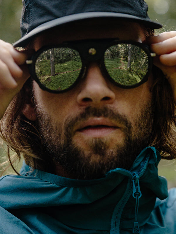 Siniseen takkiin pukeutunut henkilö säätää aurinkolaseja ja heijastaa metsäkuvaa, jossa on puita ja ruohoa.