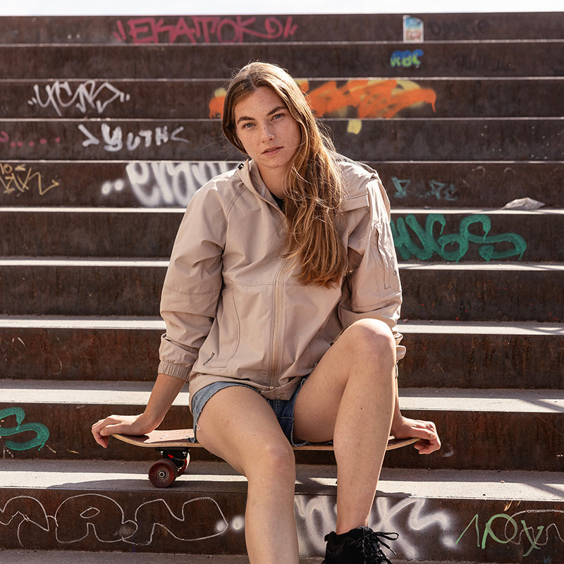 Vaaleaan takkiin ja shortseihin pukeutunut henkilö istuu portailla rullalauta kädessään värikkäiden graffitien ympäröimänä.