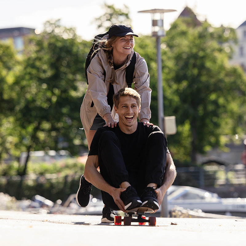 Nainen seisoo rullalaudan päällä pitäen kiinni miehen olkapäistä, kun hän istuu sen päällä, molemmat hymyilevät ulkona.