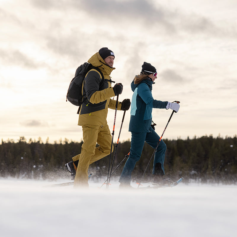 Kaksi talvivaatteisiin pukeutunutta henkilöä hiihtää lumisessa maastossa, taustalla puita.