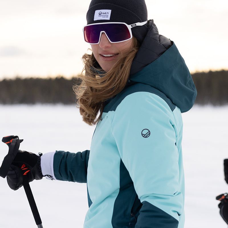 Vaaleansiniseen takkiin, mustaan pipoon ja aurinkolaseihin pukeutunut henkilö, jolla on suksisauvat lumisessa maisemassa.