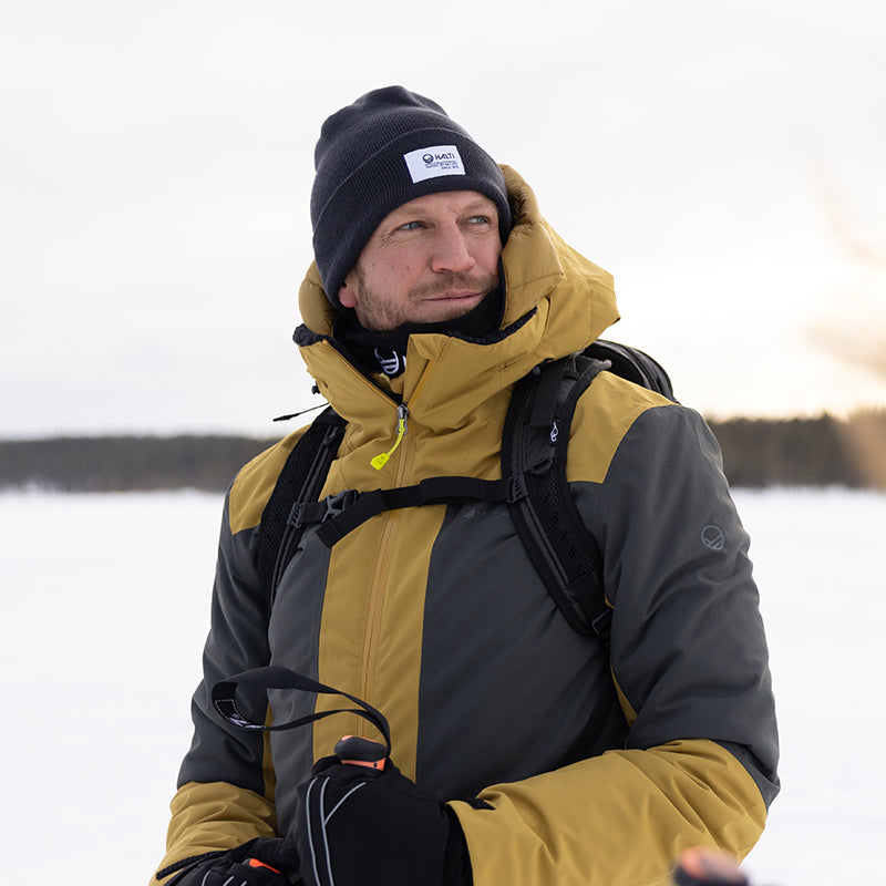 Keltaisen ja harmaan talvitakkiin ja mustaan pipoon pukeutunut mies seisoo ulkona lumisessa maisemassa suksisauvat kädessään.