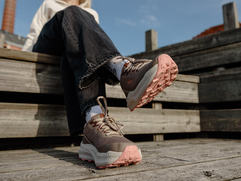 Henkilö istuu puisella penkillä, jalassa mustat farkut ja ruskeat lenkkarit vaaleanpunaisella pohjalla, jalat maassa.