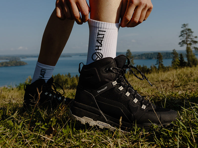 Henkilö, jolla on mustat vaelluskengät ja valkoiset sukat, säätää nauhoja ulkona, taustalla järvi ja puita.