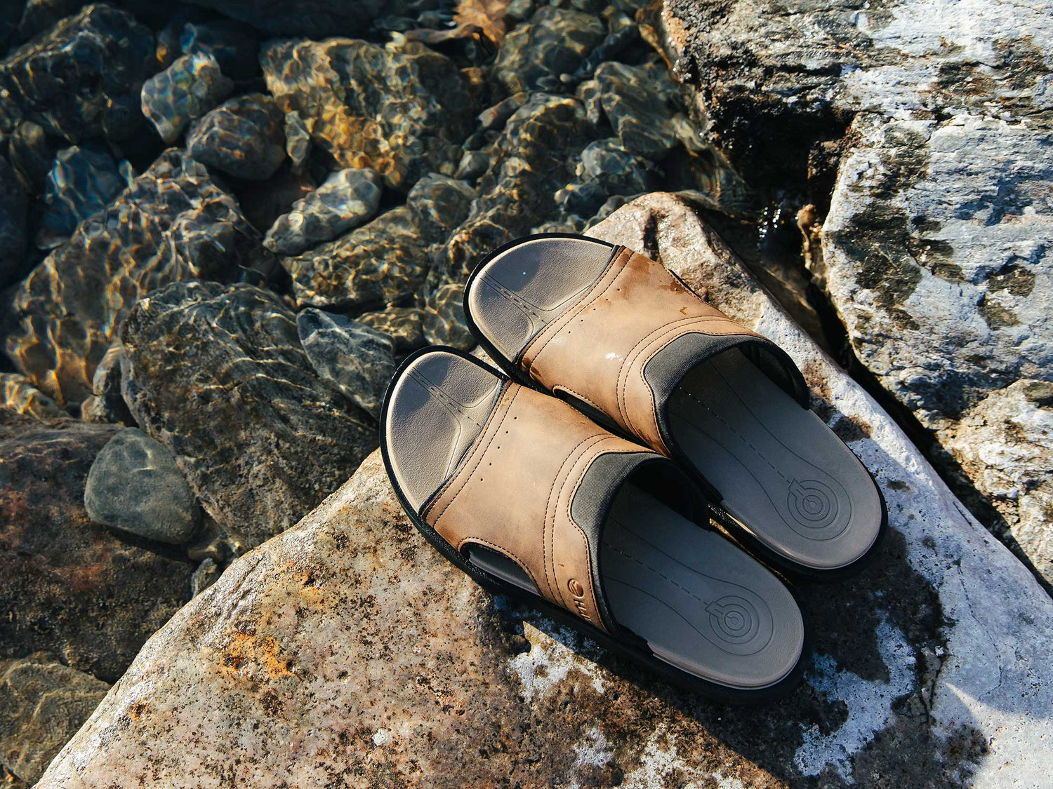 Pari ruskeita liukusandaaleja, jotka lepäävät suurella kivellä kirkkaan, matalan veden äärellä, jossa on näkyviä kiviä.