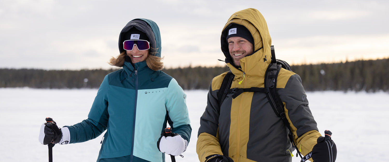 Kaksi talvitakkiin pukeutunutta ihmistä, joilla on suksisauvat kädessään, seisoo ulkona lumisessa maisemassa, jonka taustalla on puita.