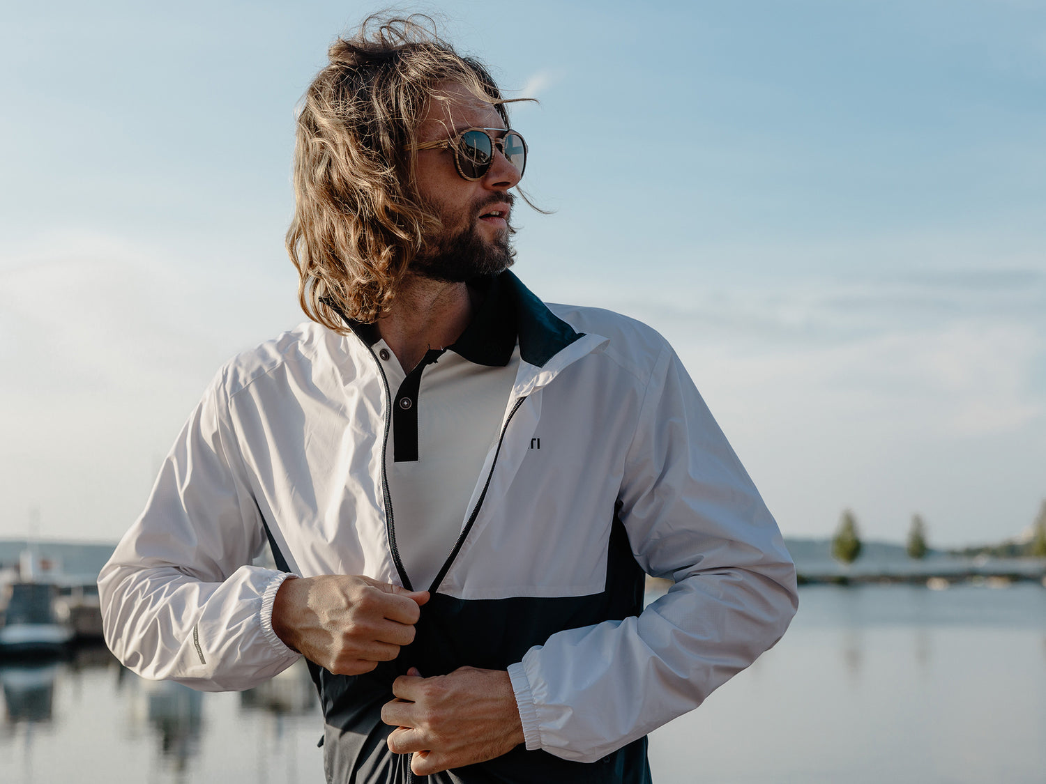 Henkilö, jolla on aurinkolasit ja takki, seisoo ulkona veden äärellä, taustalla veneitä ja puita.