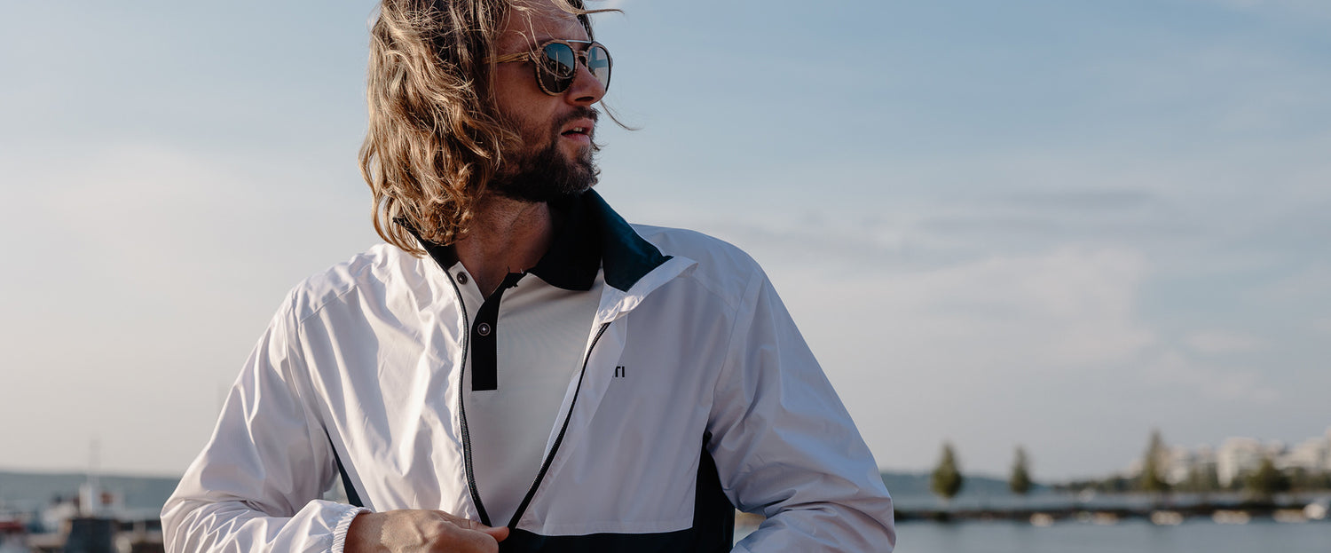 Mies, jolla on pitkät hiukset ja aurinkolasit ja valkoinen takki, seisoo ulkona veden äärellä aurinkoisena päivänä.