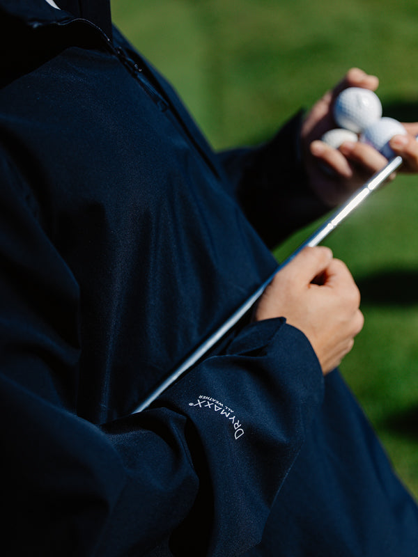 Tummiin takkeihin pukeutunut henkilö pitää golfmailaa ja kahta golfpalloa kädessään ja seisoo nurmikolla.