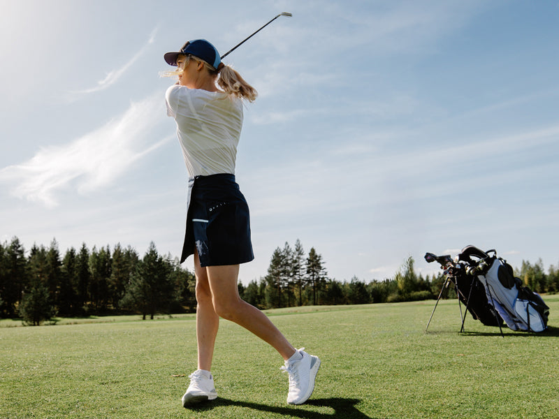 Henkilö heiluttaa golfmailaa golfkentällä, taustalla on golfbägi ja puita kirkkaalla taivaalla.