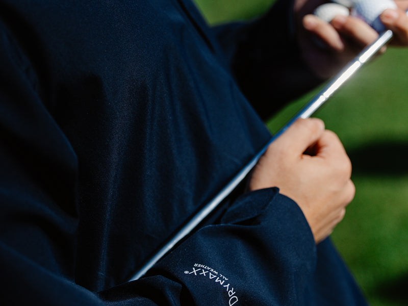 Tummassa takissa oleva henkilö pitelee golfmailaa ja golfpalloja; DRYMAXX-logo näkyy hihassa.