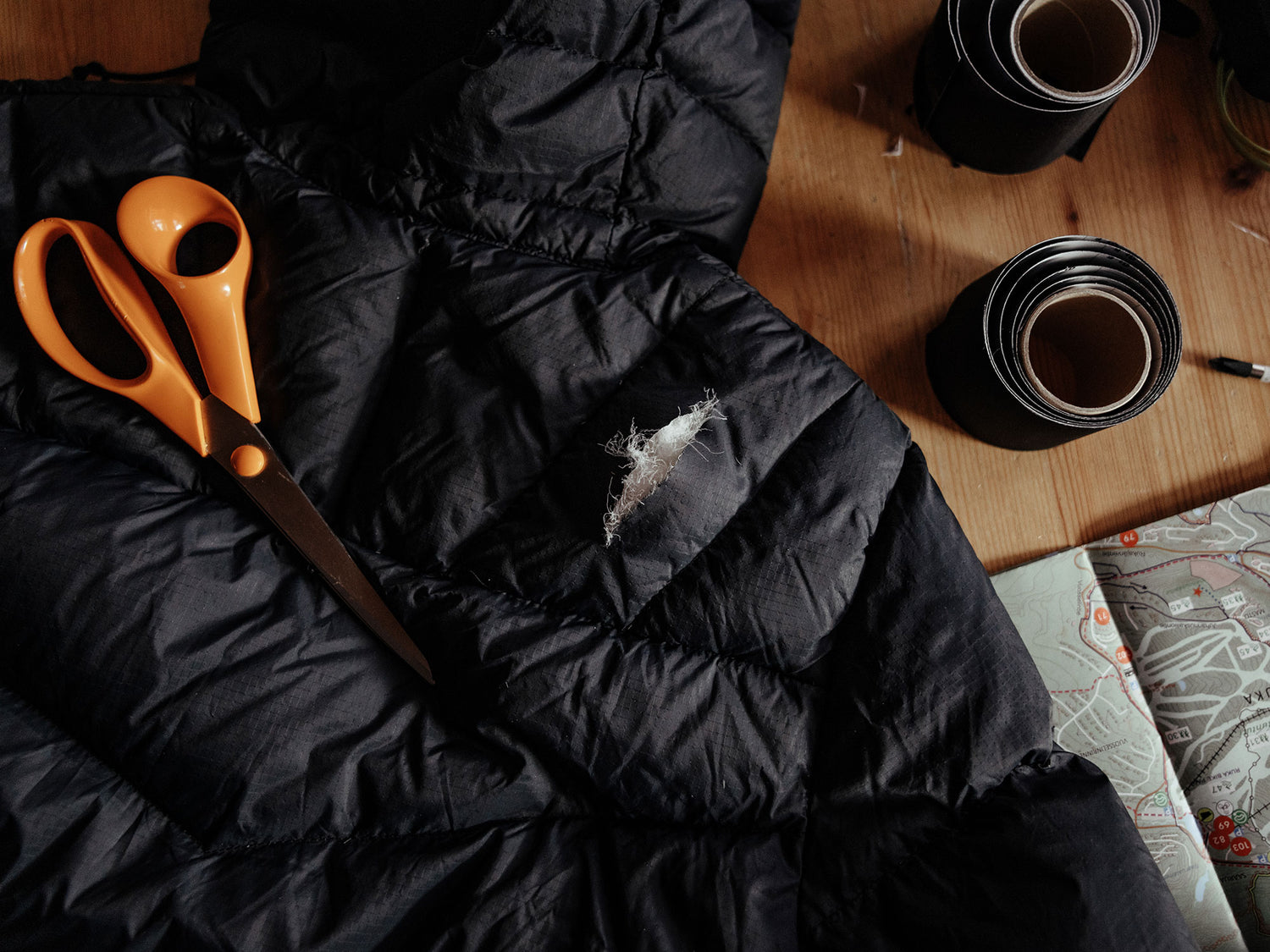 Oranssit sakset ja revitty musta takki ovat puupöydällä mustan korjausteippirullan ja kartan vieressä.