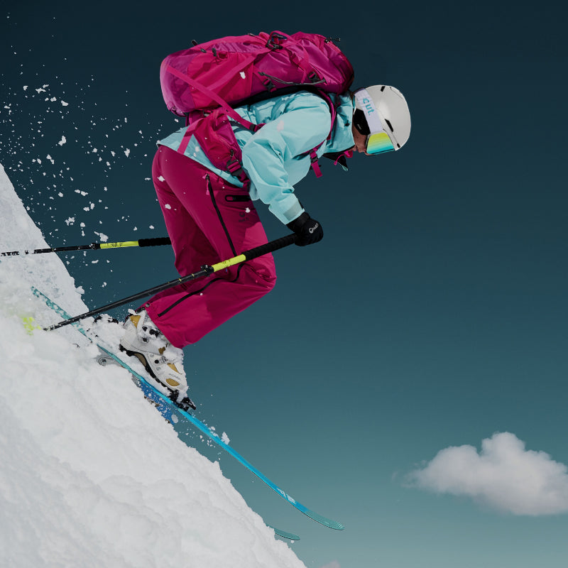 Hiihtäjä vaaleanpunaisessa ja sinisessä asussa laskeutuu lumirinteeseen sauvojen kanssa kirkkaalla taivaalla.