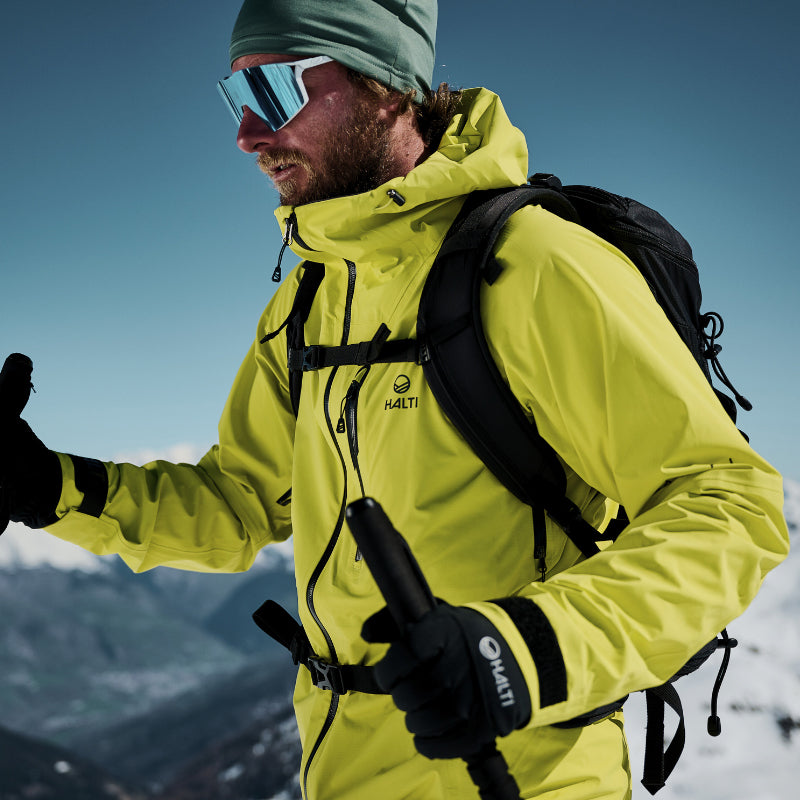 Kirkkaankeltaiseen takkiin ja aurinkolaseihin pukeutunut henkilö pitää vaellussauvoja kädessään lumisella vuorenrinteellä.