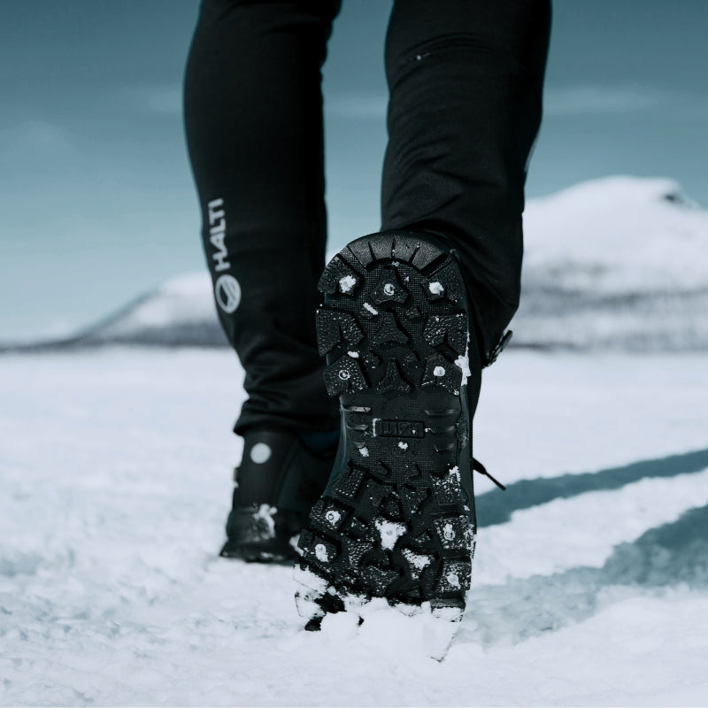 Lähikuva henkilöstä, joka kävelee lumella ja näyttää mustan saappaansa kulutuspinnan, ja jolla on mustat housut.
