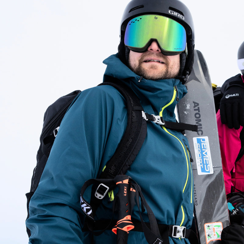 Siniseen hiihtotakkiin ja mustaan kypärään pukeutunut henkilö, jolla on sukset kädessään ja heijastavat suojalasit lumista taustaa vasten.