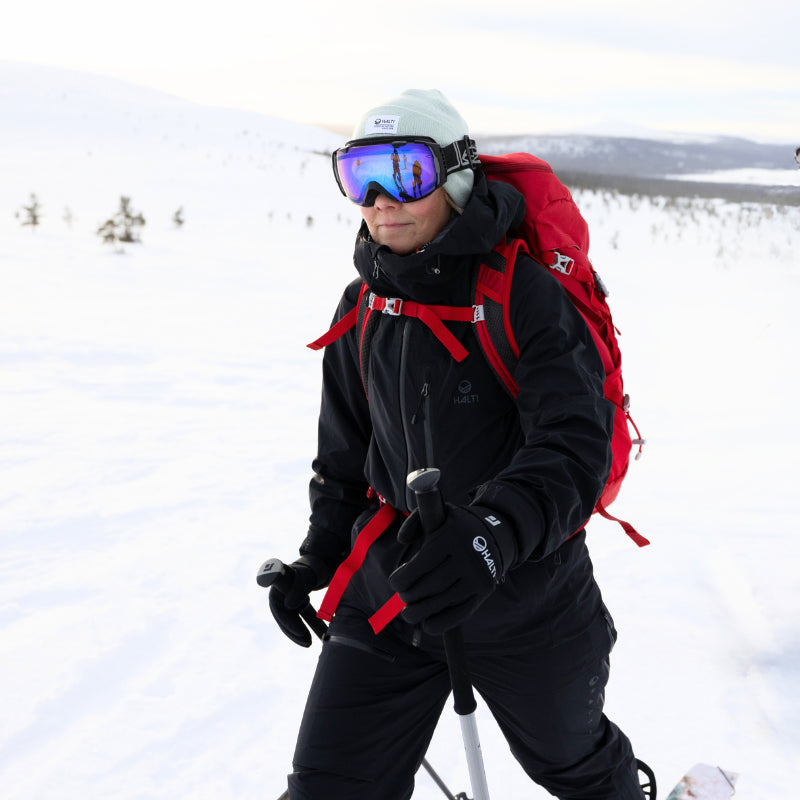 Henkilö, jolla on hiihtovarusteet ja punainen reppu, kävelee sauvojen kanssa lumisessa maisemassa, jossa on hajallaan puita.