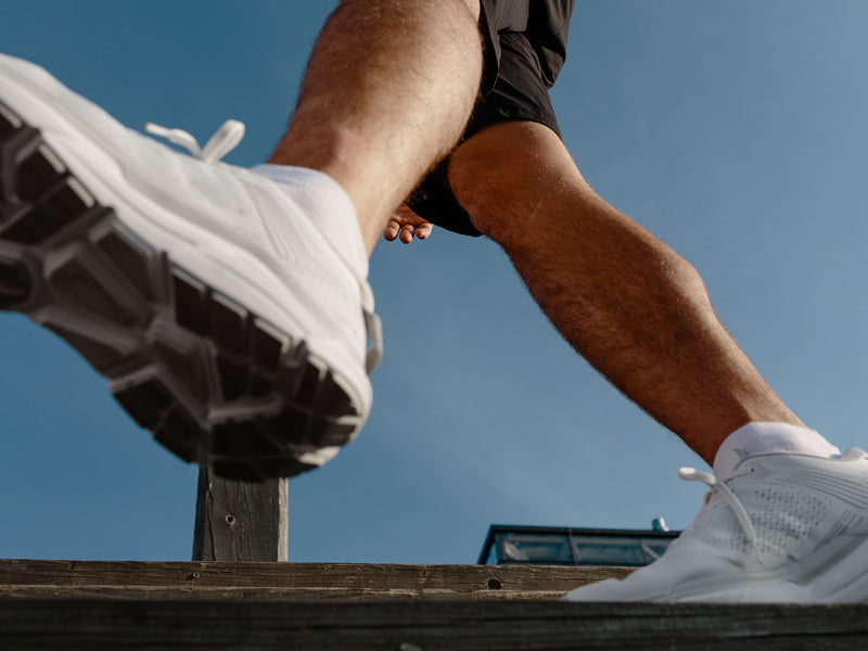 Matalakulmakuva henkilöstä, jolla on valkoiset lenkkarit ja mustat shortsit ja joka kävelee puupinnalla kirkkaalla sinisellä taivaalla.