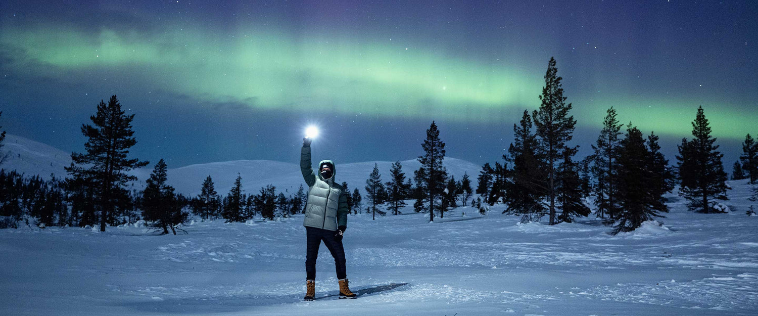 Henkilö seisoo yöllä lumisessa maastossa taskulamppu kädessään, ja yläpuolella taivaalla näkyy revontulia.