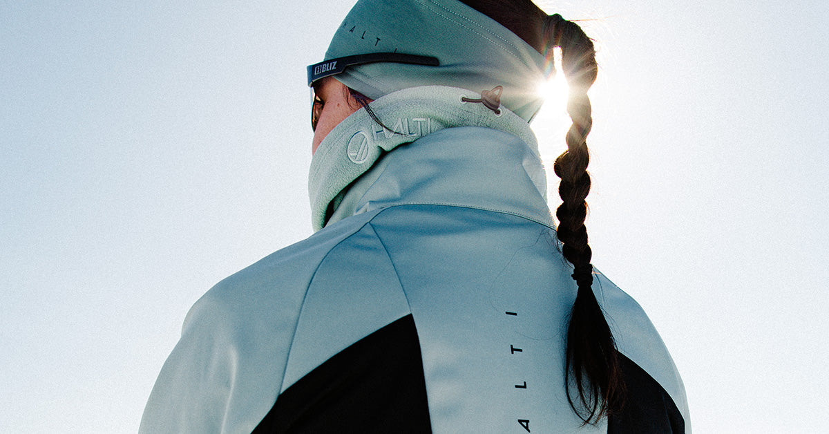 Henkilö vaaleansinisessä ja mustassa takissa, jossa on punos, takaapäin katsottuna, seisoo ulkona auringonvalon paistaessa.