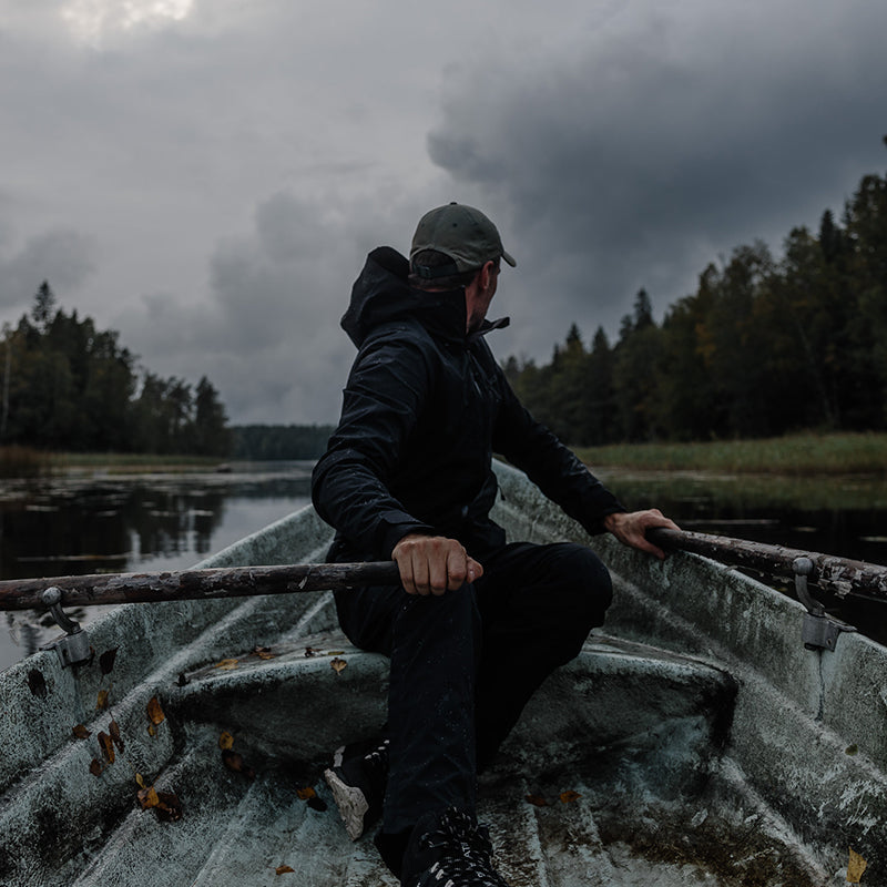 Tummiin vaatteisiin pukeutunut mies soutaa venettä rauhallisella järvellä, jota ympäröivät puut pilvisen taivaan alla.
