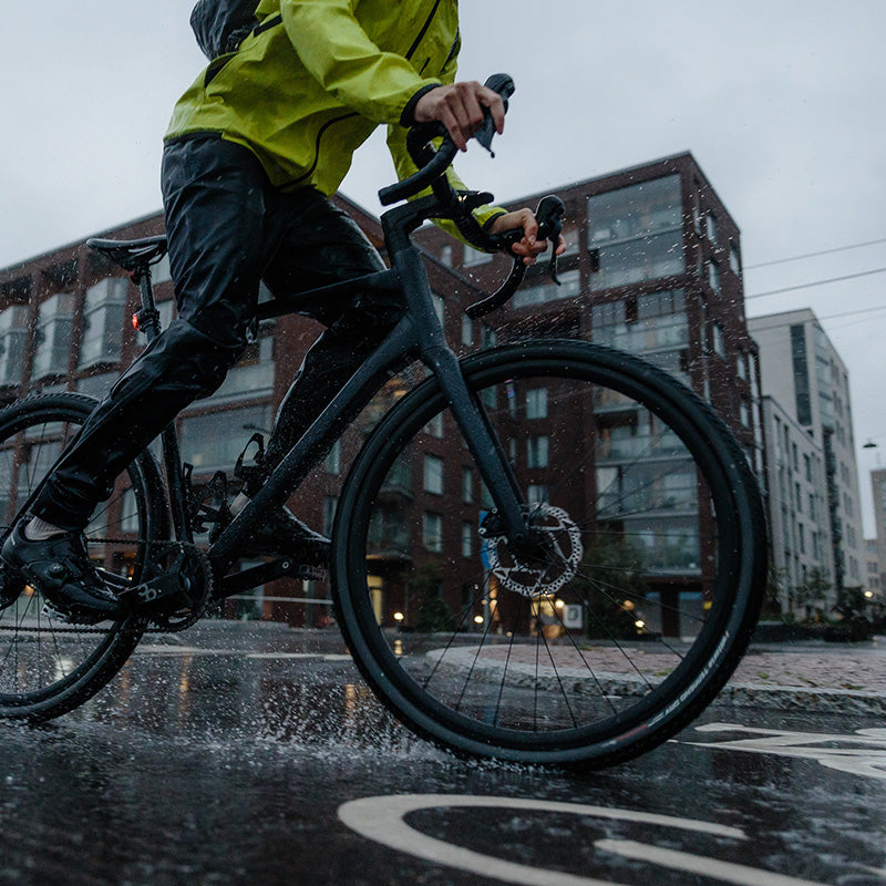 Sadevarusteisiin pukeutunut pyöräilijä ajaa polkupyörällä märällä kaupunkikadulla, ja renkaista roiskuu vettä.