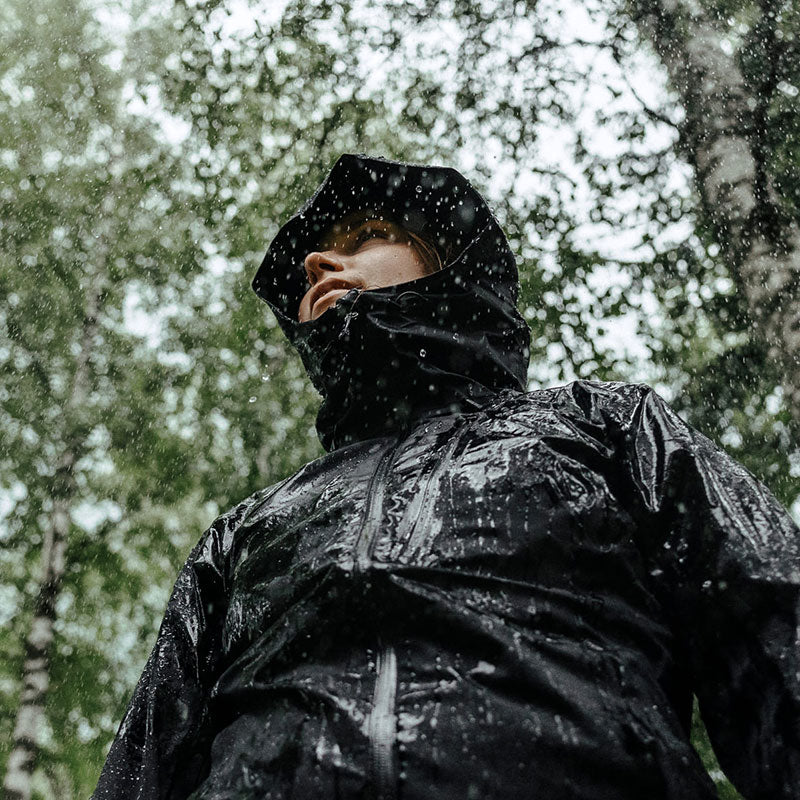 Henkilö, jolla on hupullinen sadetakki, seisoo ulkona sateessa, taustalla näkyy puita ja märkää lehdistöä.