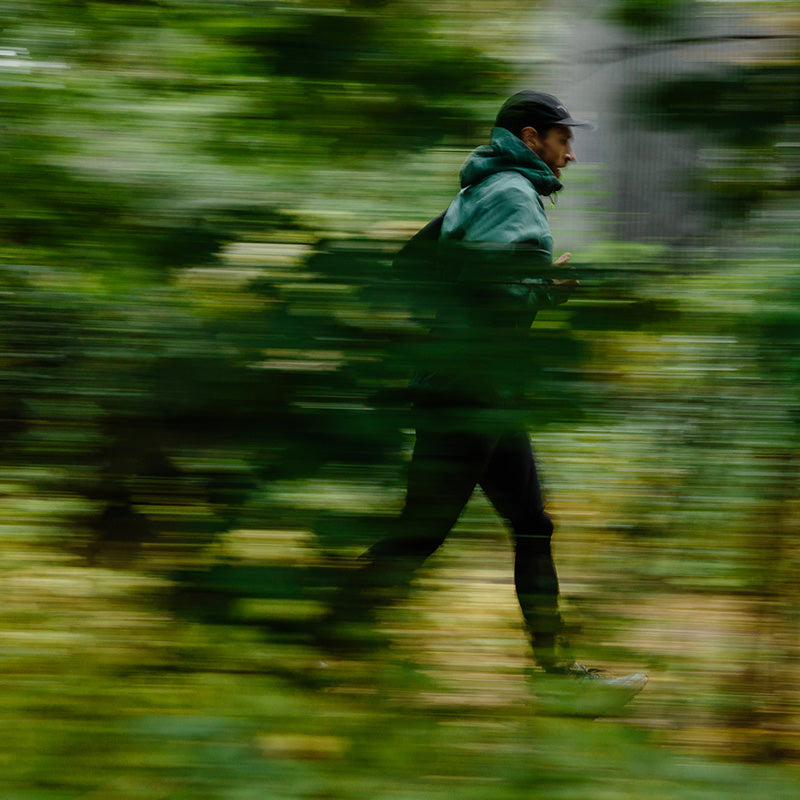 Henkilö, jolla on lippis ja takki, juoksee vihreässä, sumeassa metsäympäristössä.