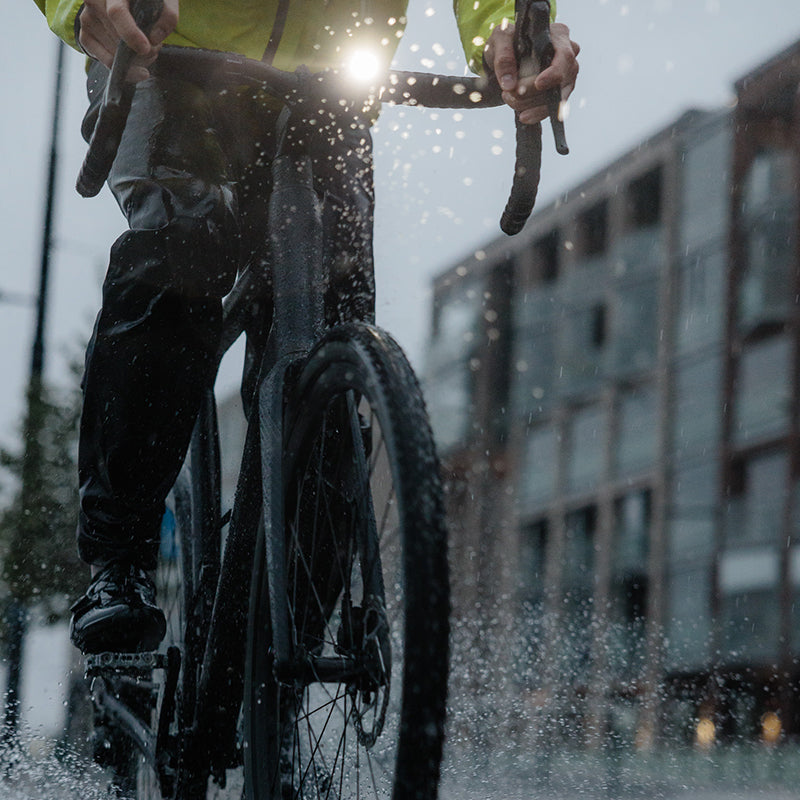 Pyöräilijä, jolla on sadevarusteet, ajaa kaupungin kadulla, ja märältä tieltä roiskuu vettä.
