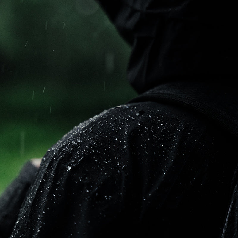 Lähikuva tummassa hupparitakissa olevasta henkilöstä, jonka olkapäällä näkyy sadepisaroita, sumea vihreä tausta.